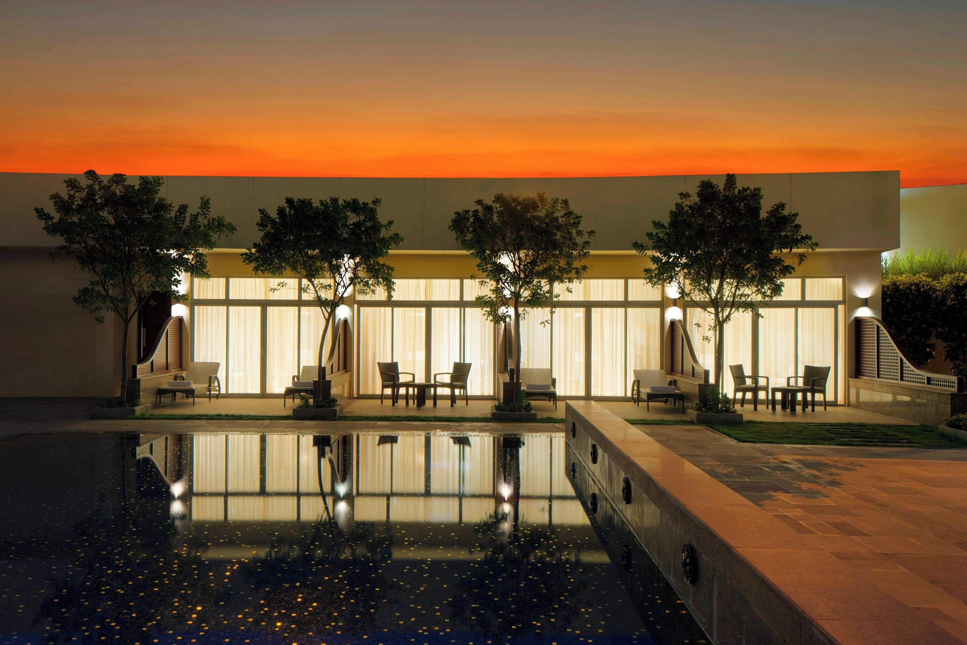 A modern building with a pool reflecting the sunset. There are outdoor seating areas with umbrellas.