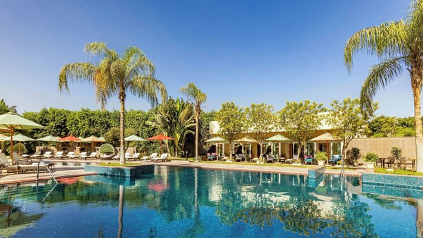 Una piscina con sillones de descanso y sombrillas, rodeada de palmeras y follaje.