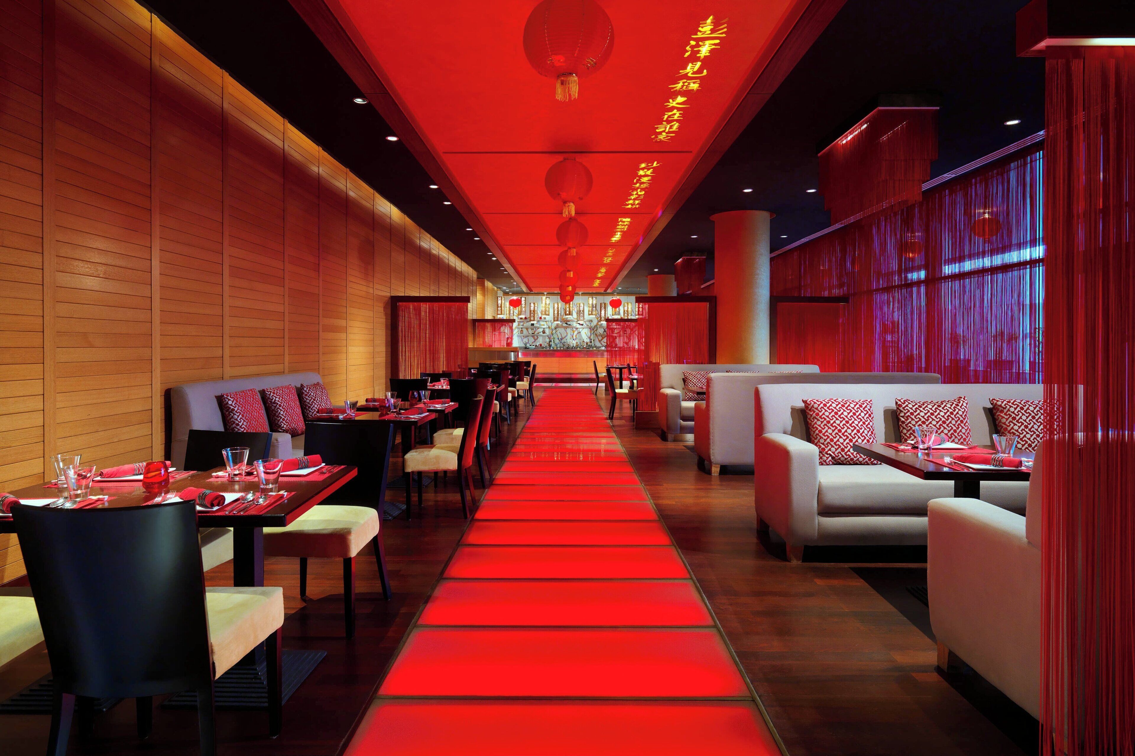 A restaurant with a red carpet leading to a central dining area, wooden panel walls, and red lanterns.