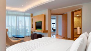 Habitación de hotel con una cama grande, televisión montada en la pared, zona de estar con una silla y una mesita, y vistas por la ventana.