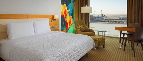 A hotel room with a large bed, a desk, a chair, and a view of an airport.