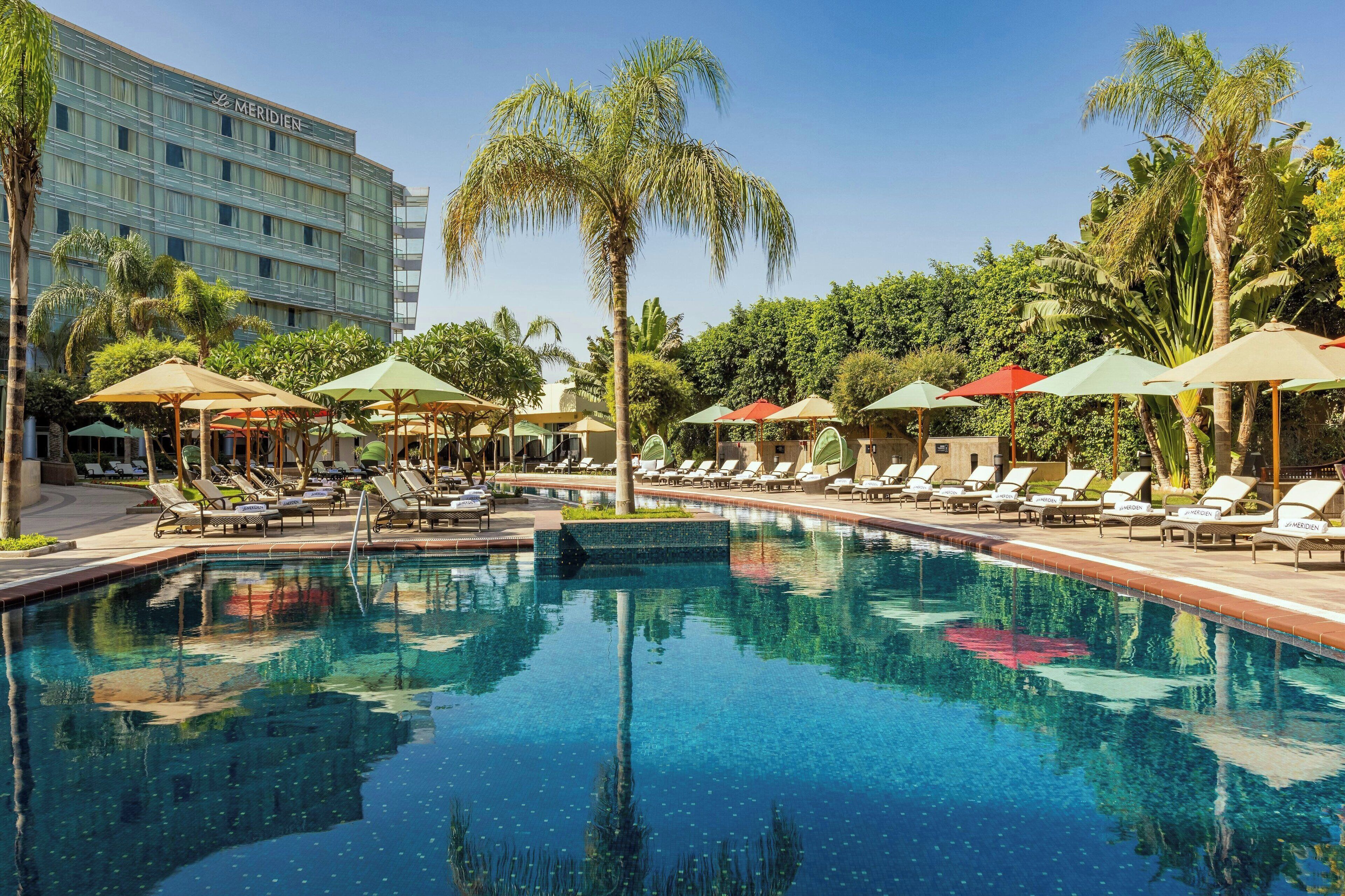 Una piscina con sillas de descanso y sombrillas, rodeada de palmeras y un edificio de hotel.