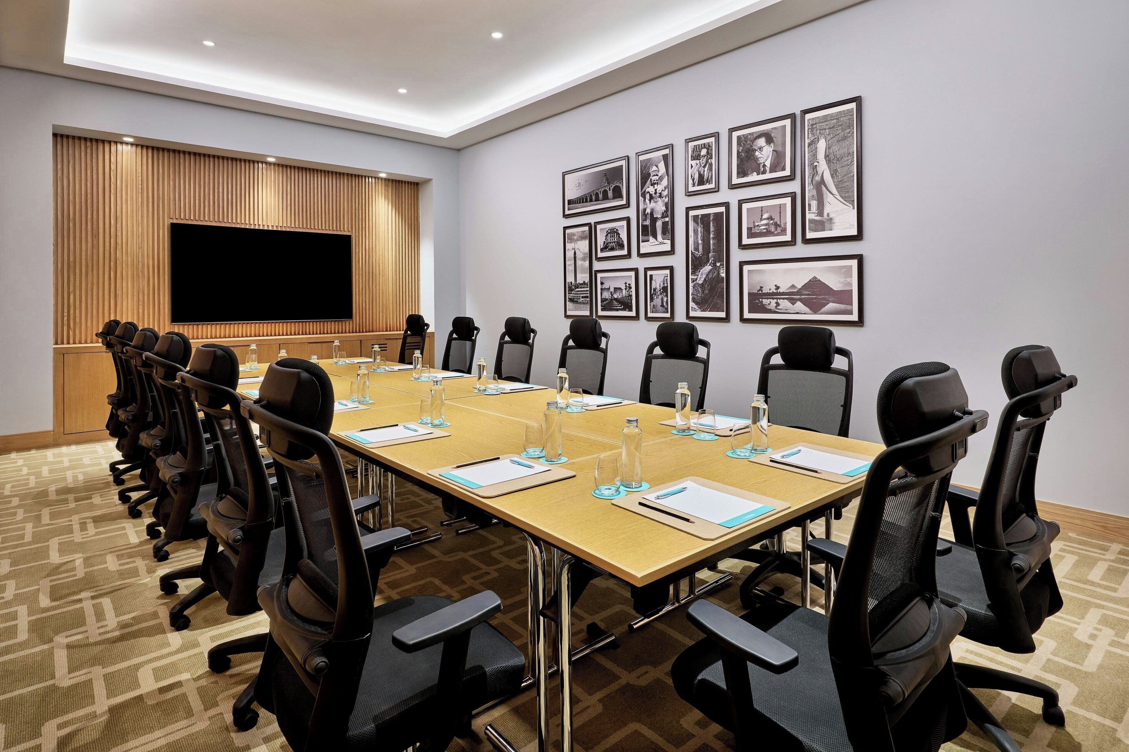 Un salón de conferencias con una larga mesa, sillas, una pantalla grande y cuadros en la pared.
