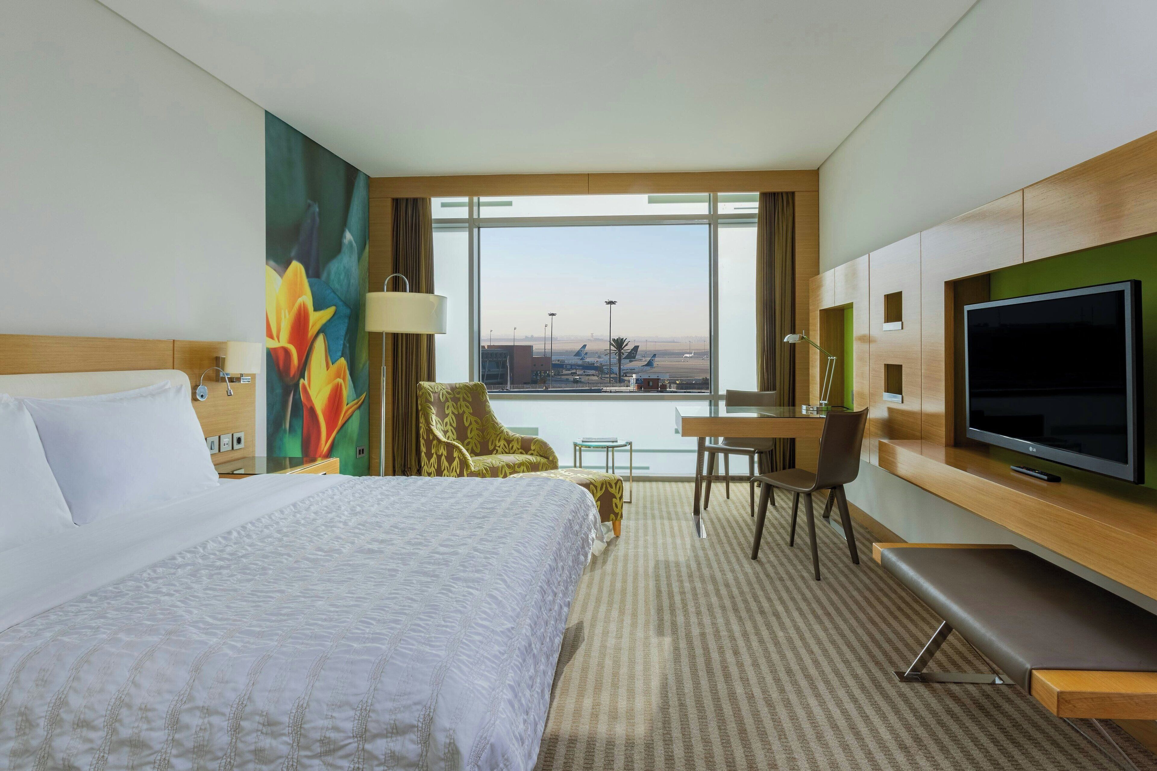 Habitación de hotel con una cama grande, un escritorio con silla, televisión y vista al aeropuerto.