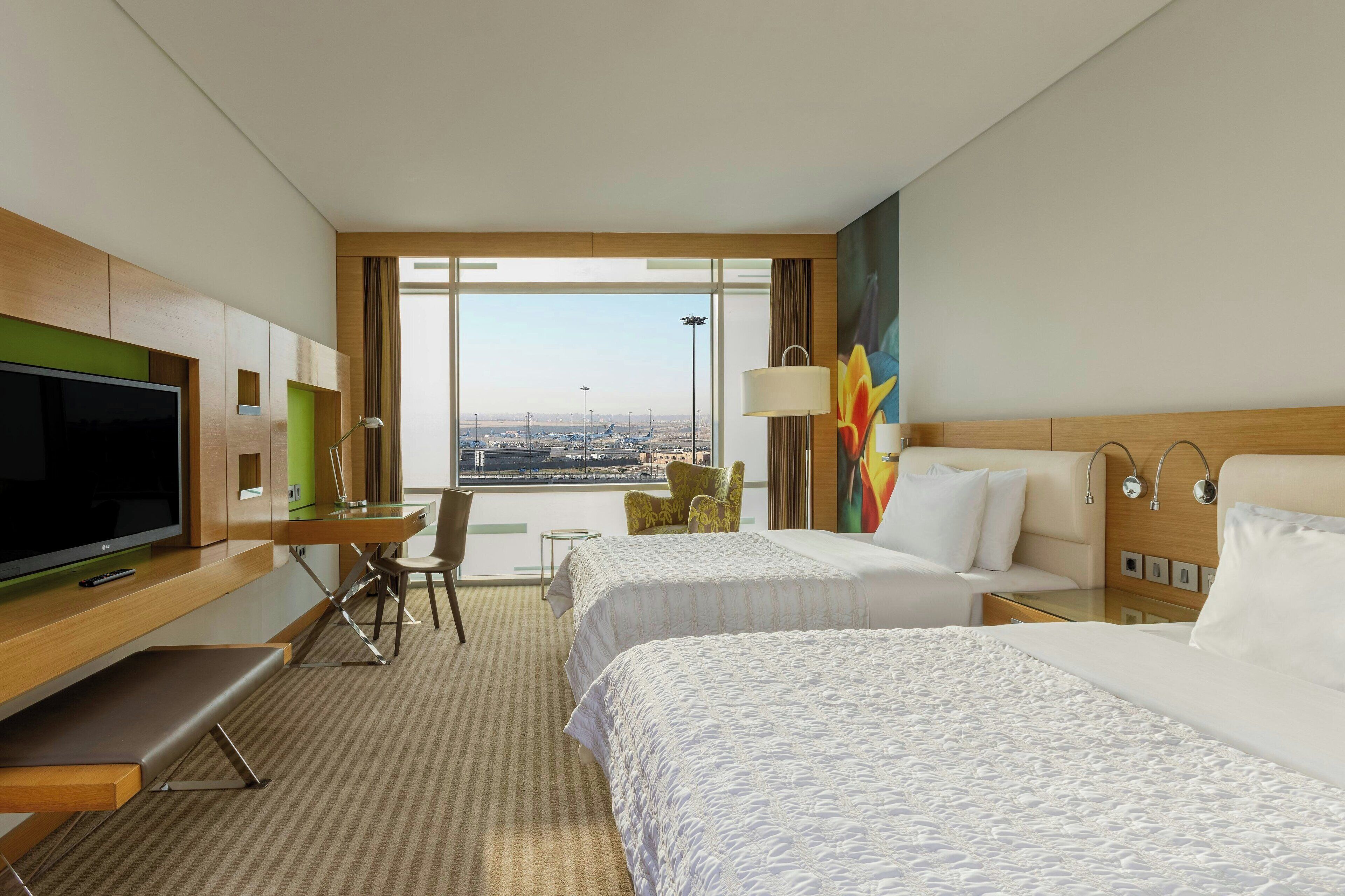 A hotel room with two beds, a desk with a TV, a chair, and a large window offering a city view.