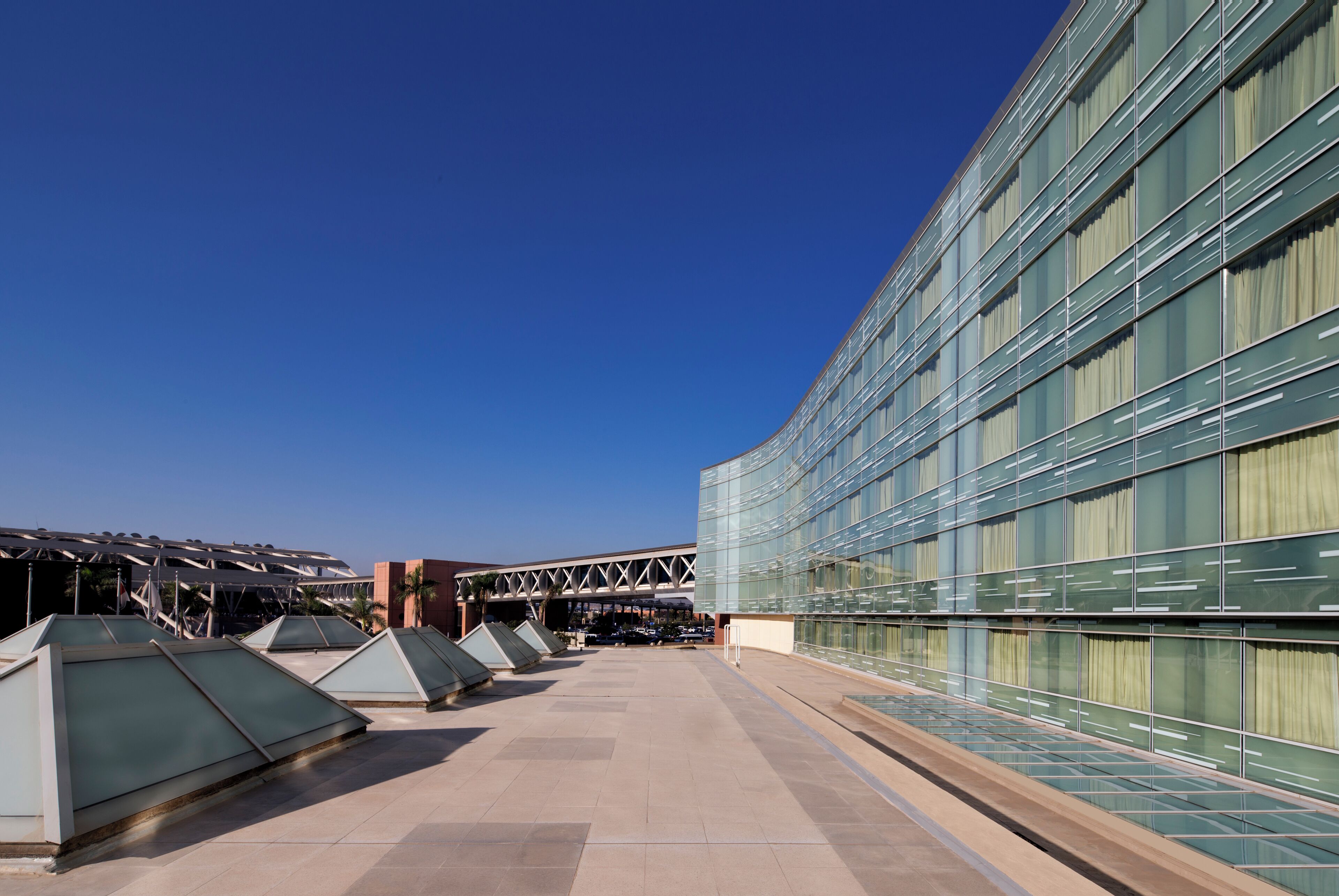 A modern building with a glass facade, a covered outdoor area with geometric structures, and a clear blue sky.