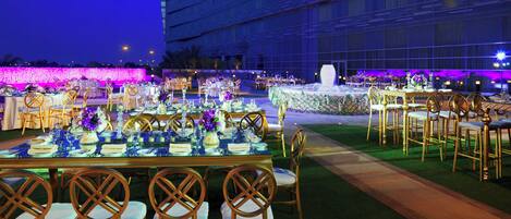 Uma área de refeições ao ar livre com mesas preparadas para um evento formal, cercada por um edifício moderno com uma placa visível.