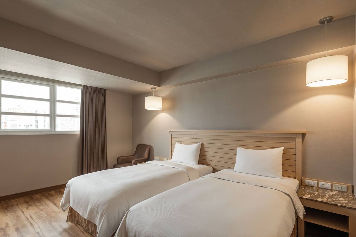 Une chambre d’hôtel avec deux lits, une chaise, une fenêtre avec des rideaux et une lampe fixée au mur.