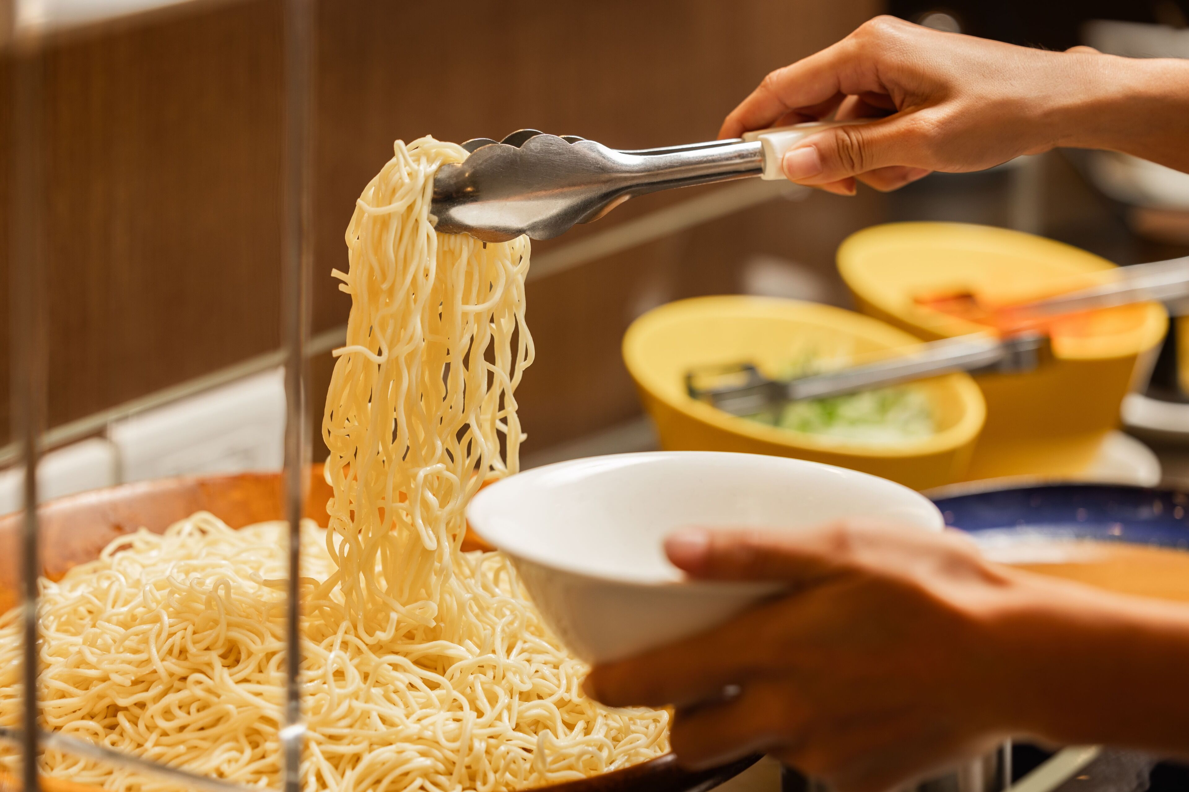 A person using tongs to serve noodles from a large pot into a bowl.