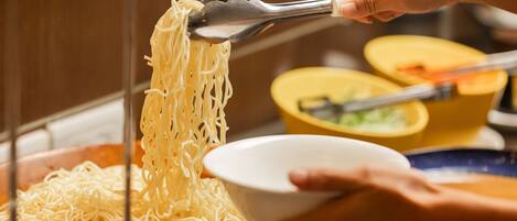 A person using tongs to serve noodles from a large pot into a bowl.