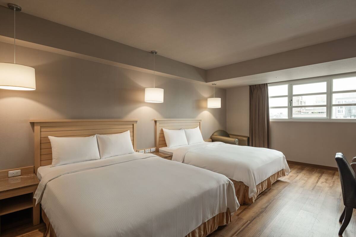 Une chambre d’hôtel avec deux lits, des têtes de lit en bois, des tables de chevet, un bureau et une fenêtre donnant sur des immeubles.