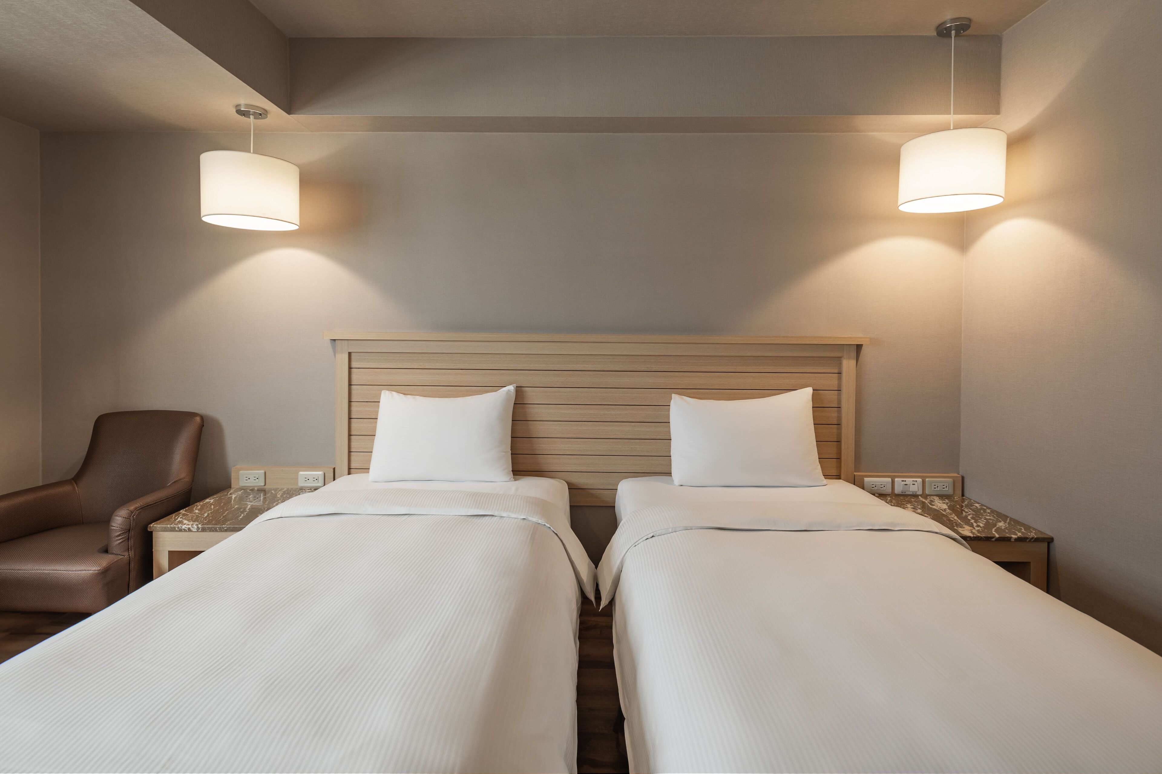 A hotel room with two beds, a wooden headboard, a brown leather chair, and two bedside tables with lamps.