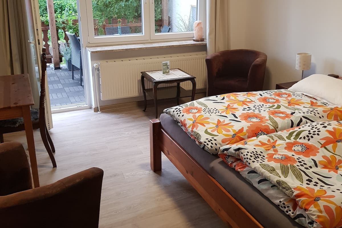 Een slaapkamer met een houten bed, een klein tafeltje, een stoel en uitzicht op een tuin door een raam.