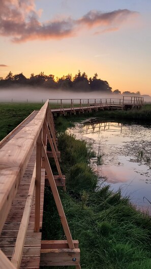 Un puente de madera cruzando un cuerpo de agua tranquilo, con un amanecer o atardecer al fondo.