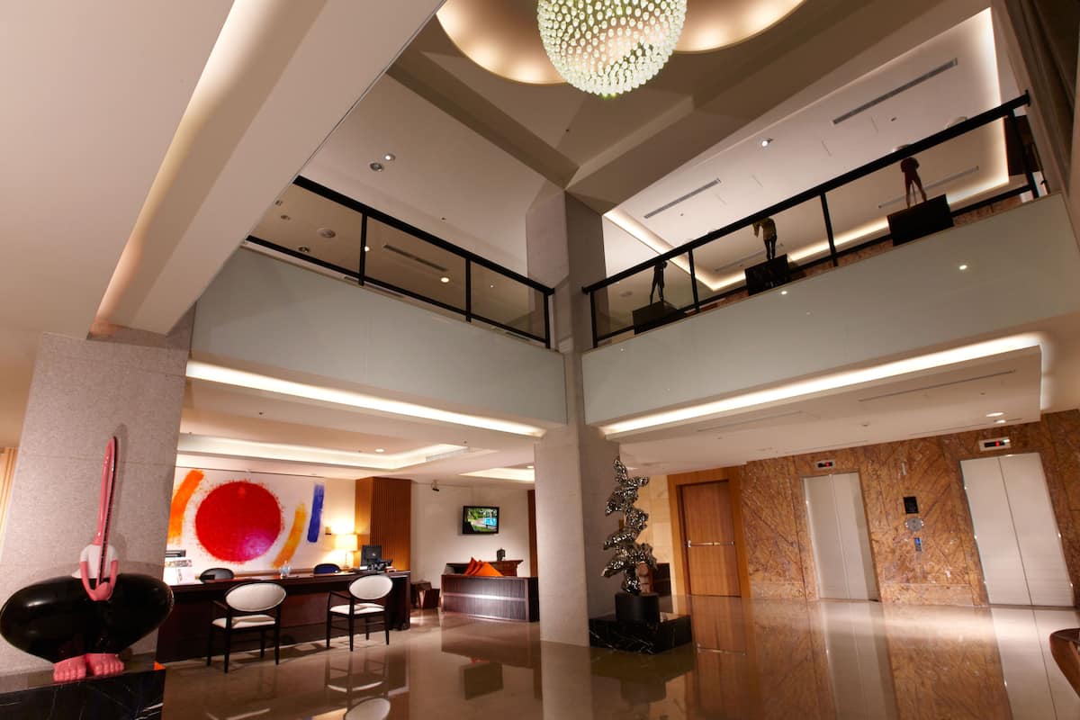 A modern hotel lobby with a high ceiling, a chandelier, a reception desk, a TV, and a sculpture.