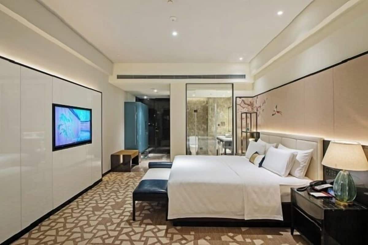 A modern hotel room with a large bed, a bedside table with a lamp, a TV mounted on the wall, and a patterned floor.