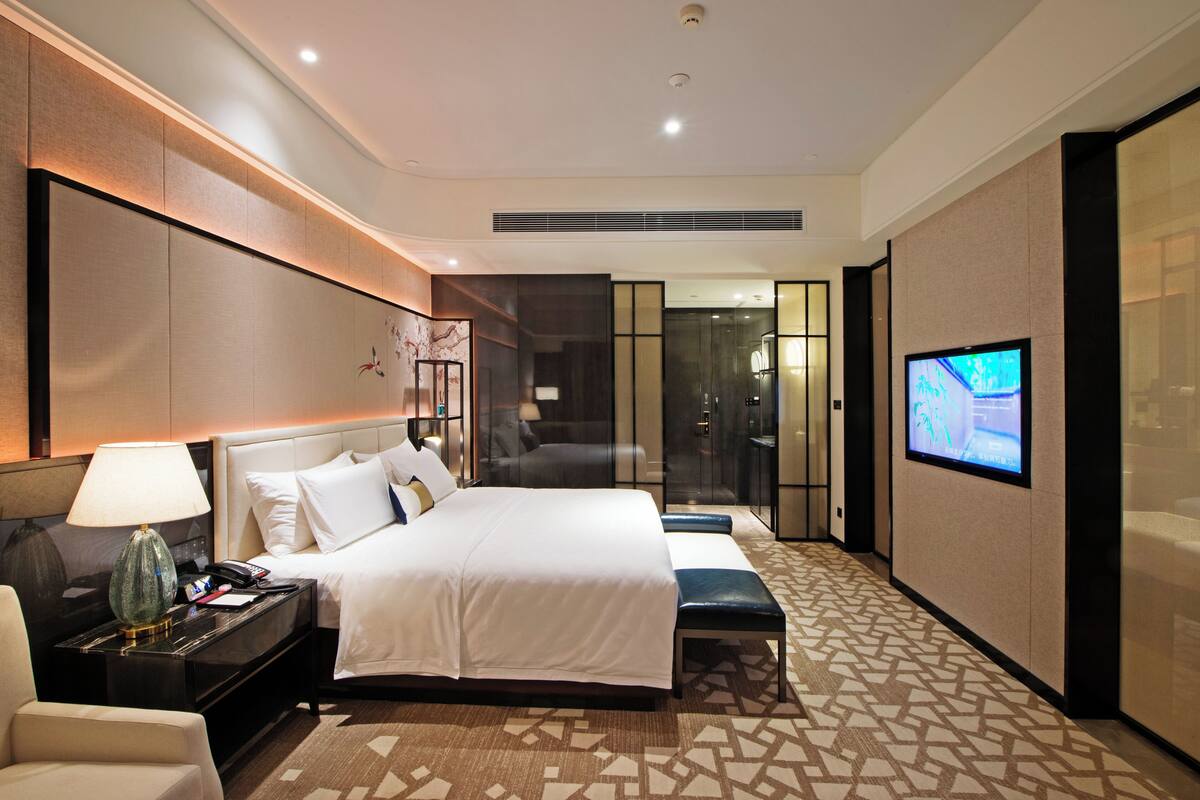 A hotel room with a large bed, bedside tables, a TV, and a bathroom visible through a glass door.