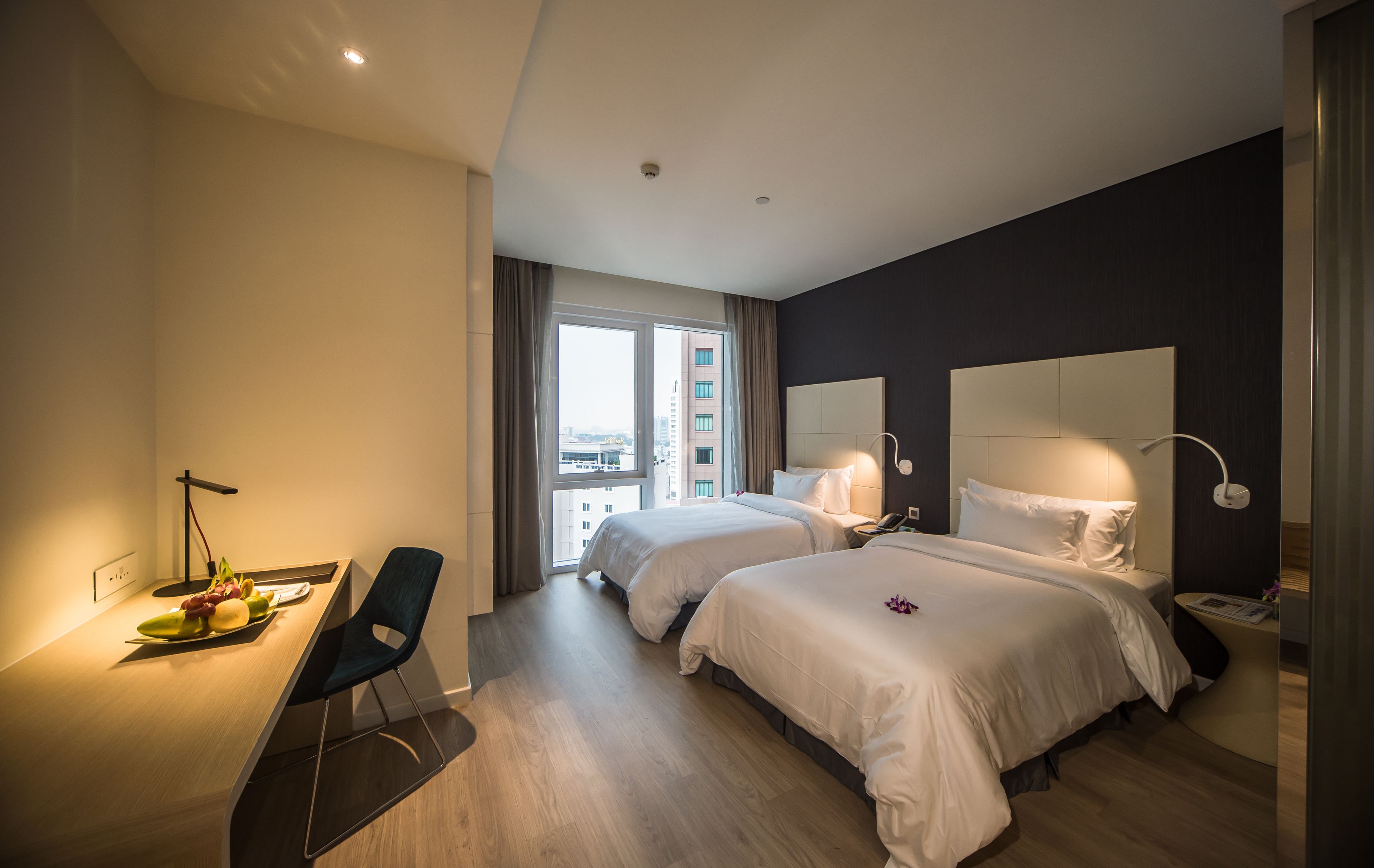 Een moderne hotelkamer met twee bedden, een bureau met stoel, een groot raam en uitzicht op de stad.