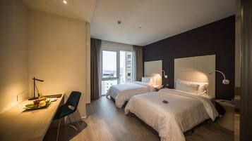 Een moderne hotelkamer met twee bedden, een bureau met stoel, een groot raam en uitzicht op de stad.