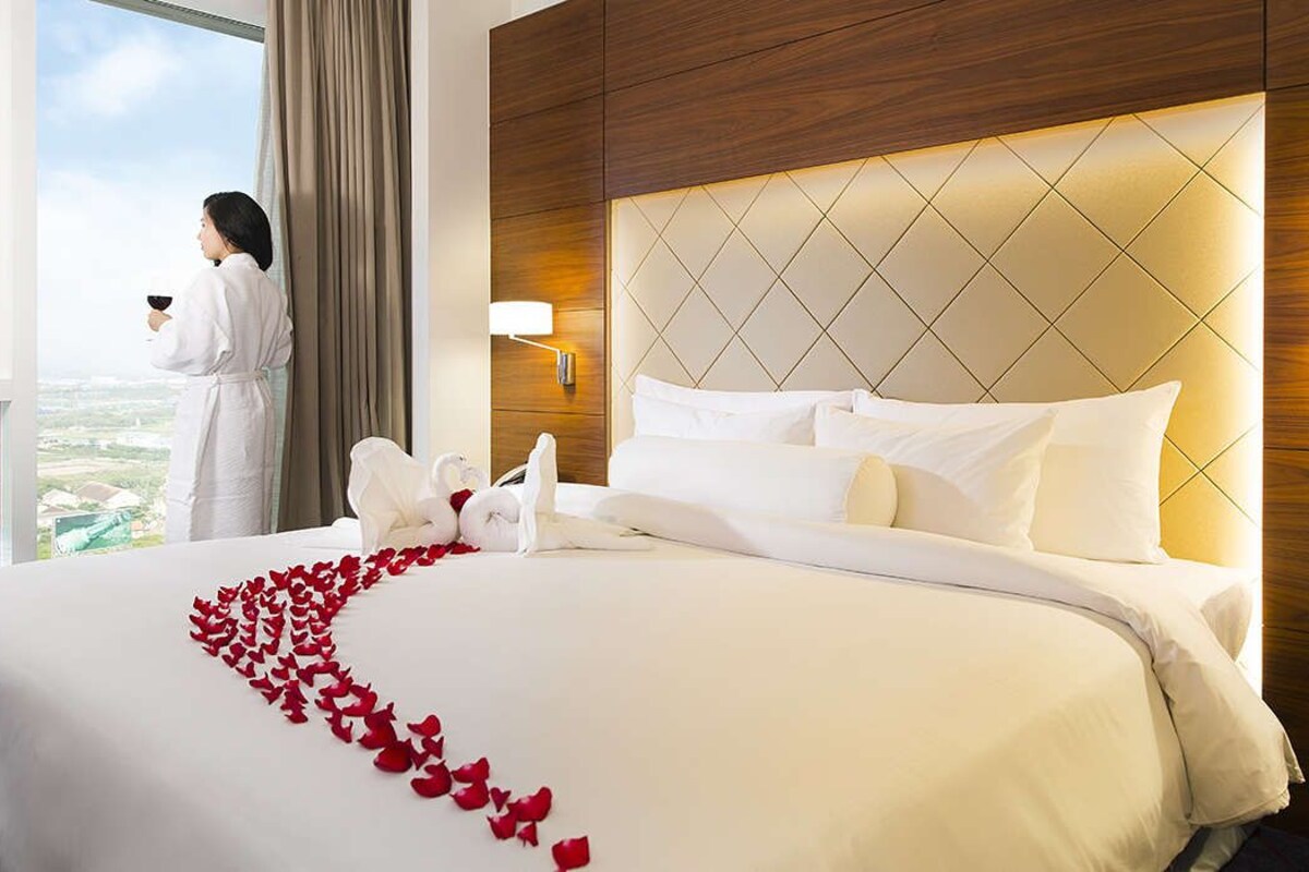 Habitación de hotel con una cama grande, vistas a la ciudad y una persona con bata junto a la ventana.
