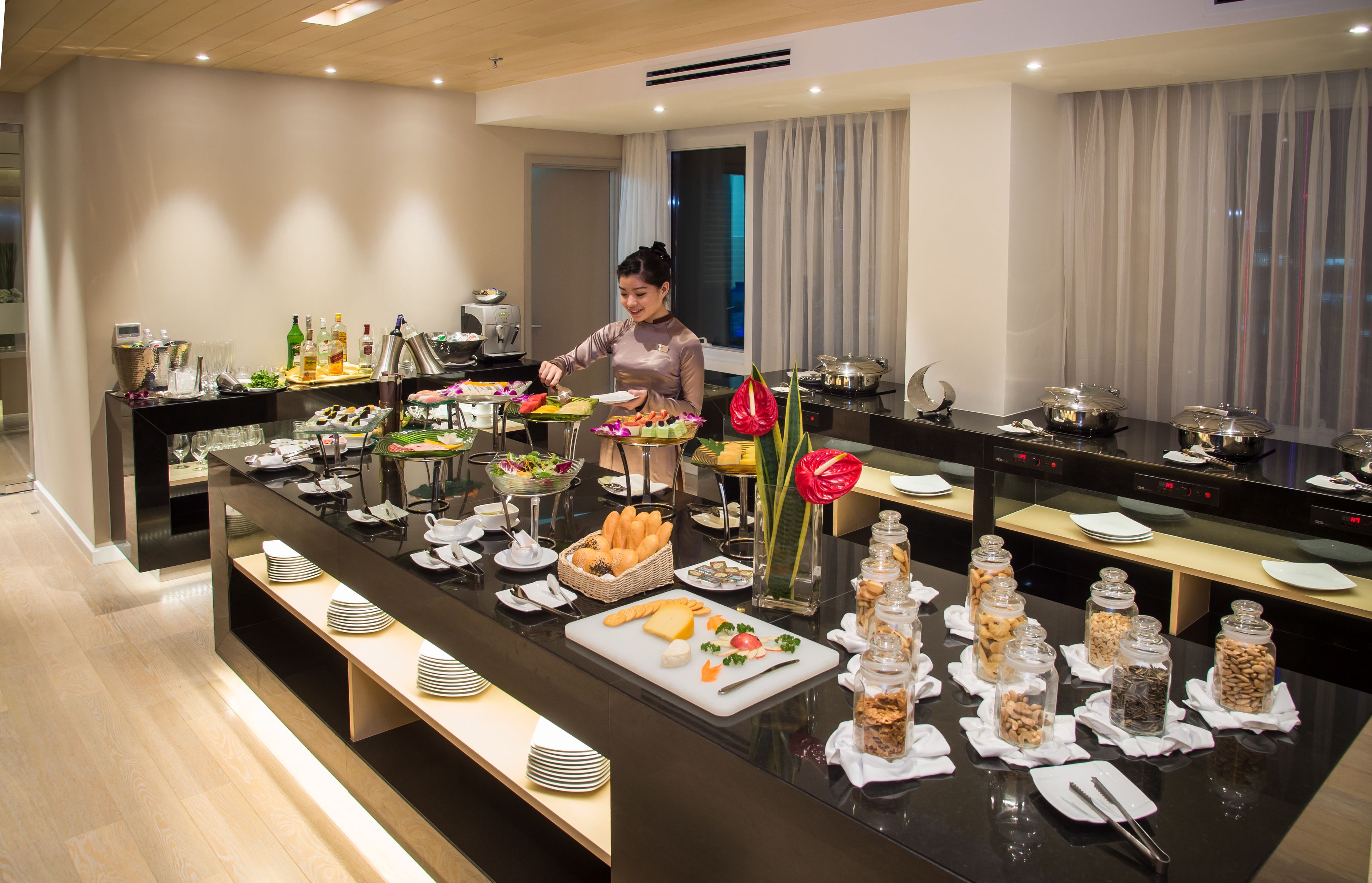 Montaje de bufé con diversos alimentos y salsas, un encargado de bufé y un interior moderno con un ventanal grande y cortinas.