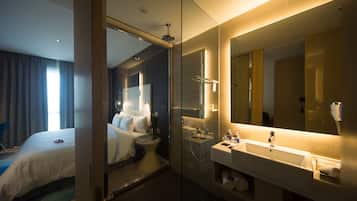 Ein modernes Hotelzimmer mit einer gläsernen Dusche, einem großen Spiegel, einem weißen Waschbecken und einem ordentlich bezogenen Bett.