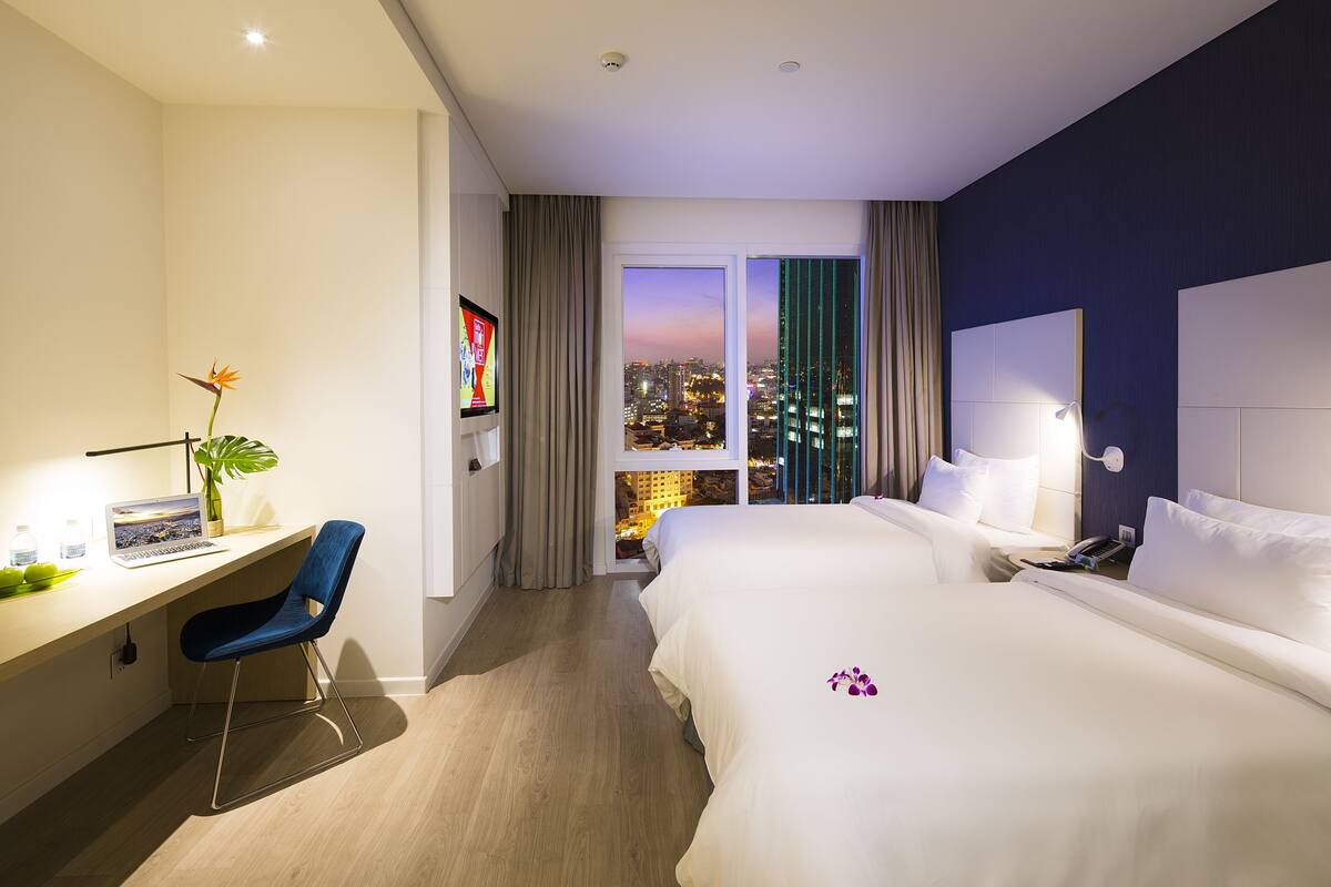 Quarto de hotel com escrivaninha, duas camas, vista para a cidade e uma planta.