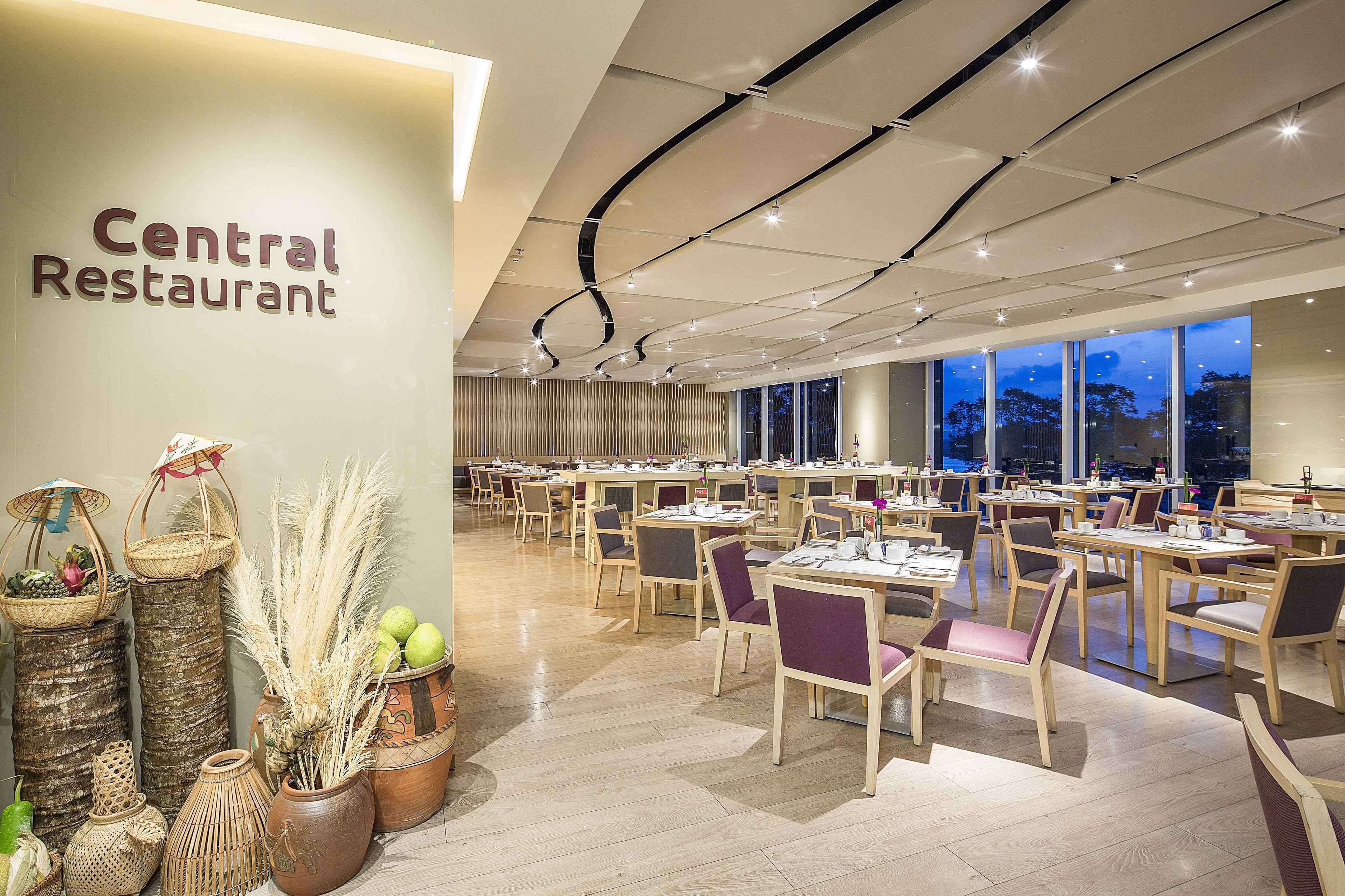 A spacious restaurant with a modern interior, featuring a central wall with the restaurant's name, numerous tables and chairs, and large windows offering a view of the outside.