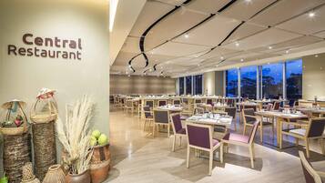 A spacious restaurant with a modern interior, featuring a central wall with the restaurant's name, numerous tables and chairs, and large windows offering a view of the outside.