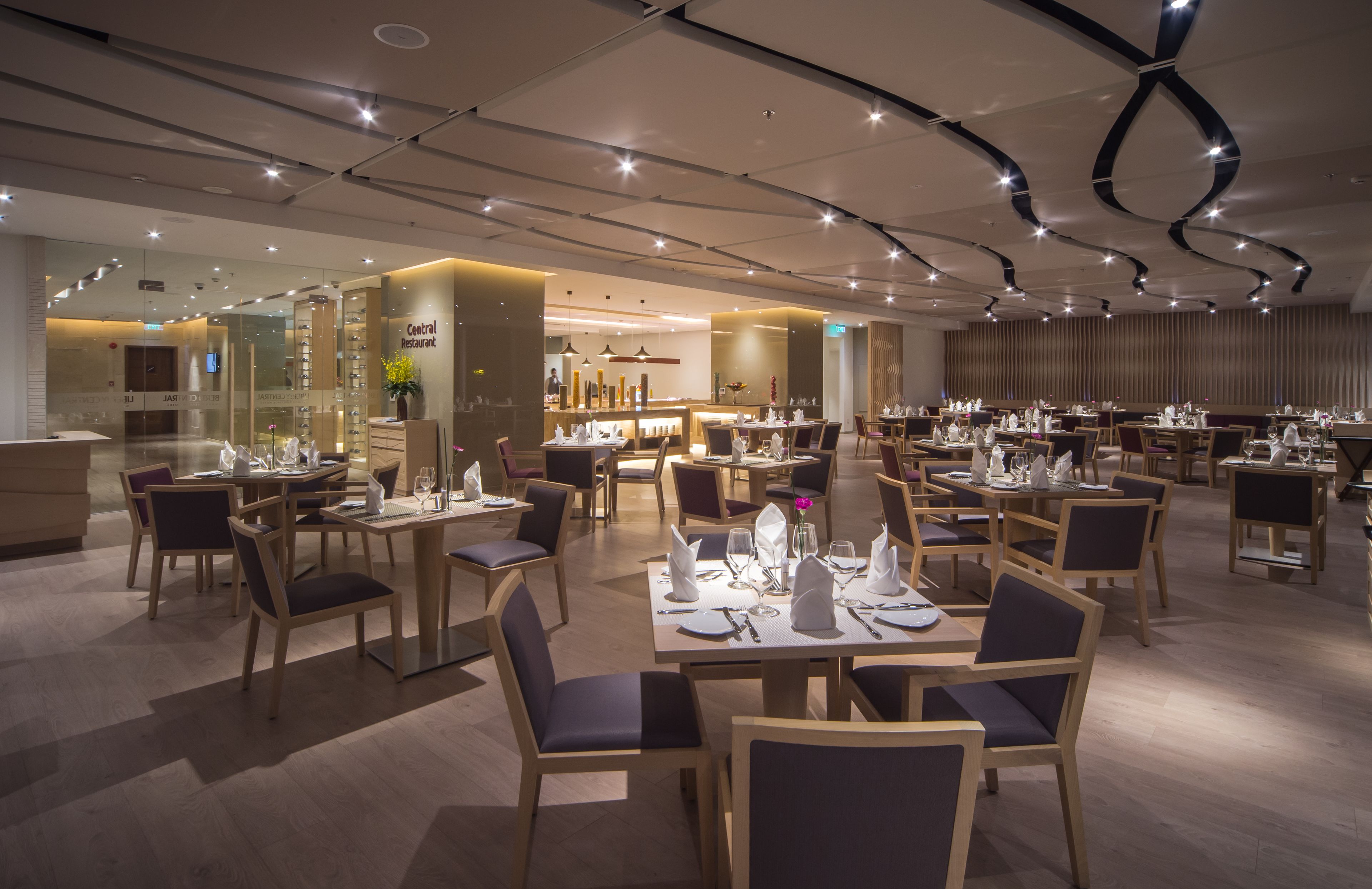 A modern restaurant with multiple dining tables and chairs, a central bar area, and a well-lit ceiling with unique lighting fixtures.