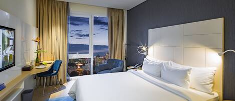 Une chambre d’hôtel avec un grand lit, un bureau avec une chaise, une télévision et une vue sur la ville la nuit.