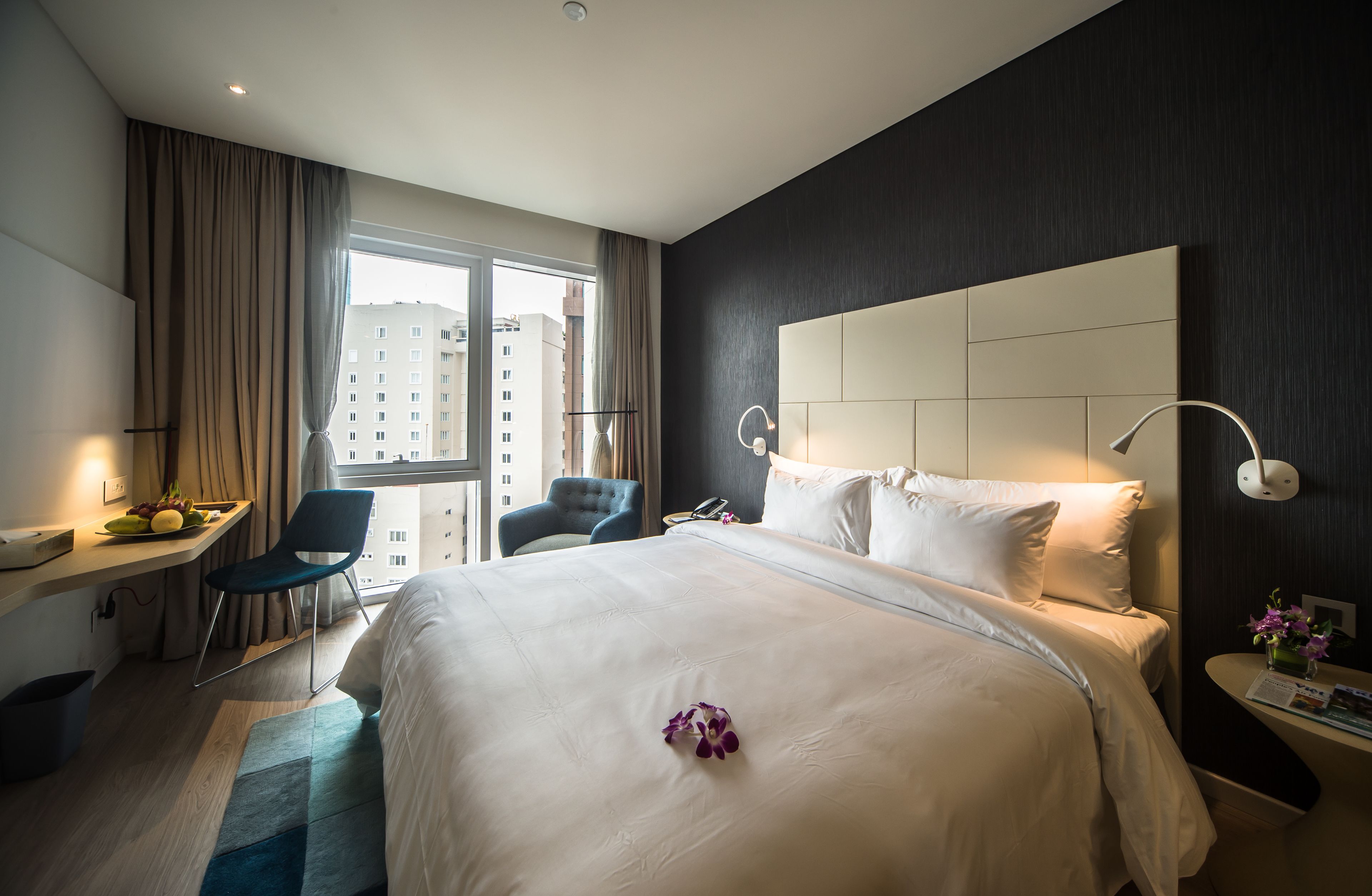 Ein modernes Hotelzimmer mit einem großen Bett, einem Schreibtisch mit Stuhl, einem Nachttisch mit Lampe und Blick auf die Stadt durch ein großes Fenster.