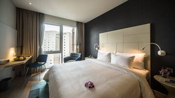 Ein modernes Hotelzimmer mit einem großen Bett, einem Schreibtisch mit Stuhl, einem Nachttisch mit Lampe und Blick auf die Stadt durch ein großes Fenster.