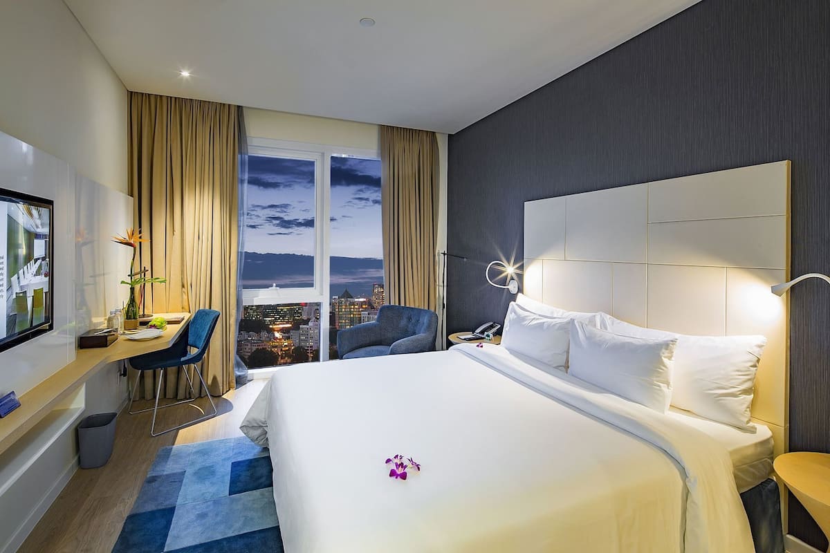 Quarto de hotel com uma cama grande, uma escrivaninha com cadeira, televisão e vista para a cidade pela janela.