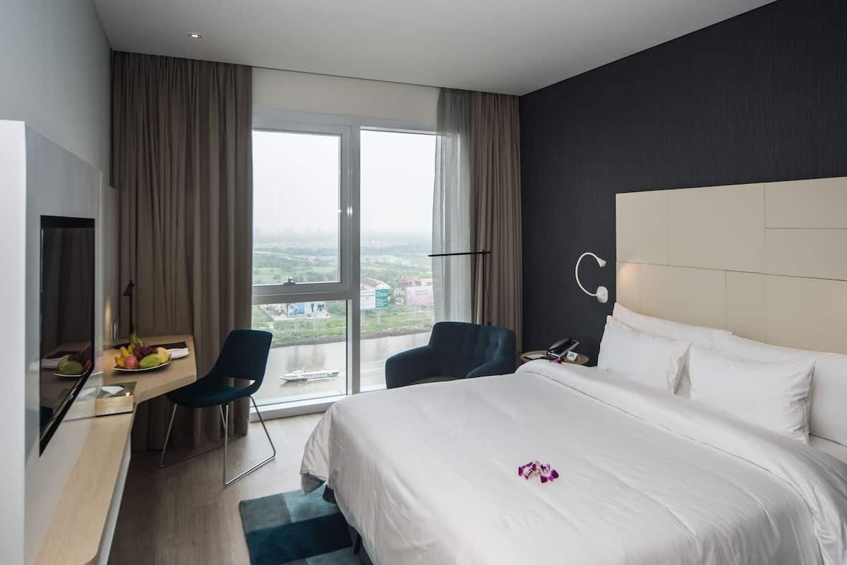 Quarto de hotel moderno com uma cama grande, uma escrivaninha com cadeira, vista para a cidade pela janela e uma mesa de cabeceira com abajur.