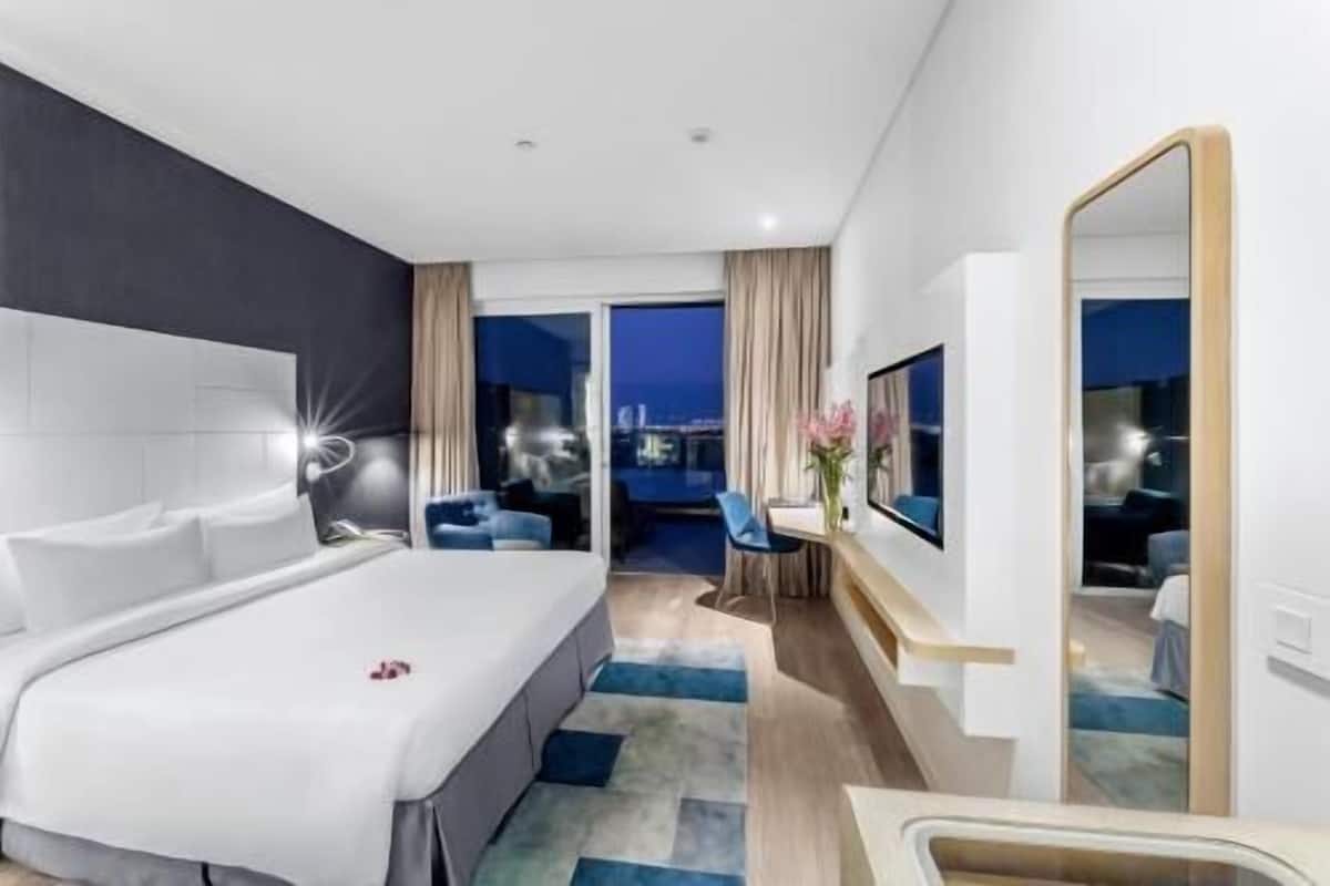 Quarto de hotel moderno com uma cama grande, uma escrivaninha, uma cadeira, um espelho e vista para a cidade à noite.