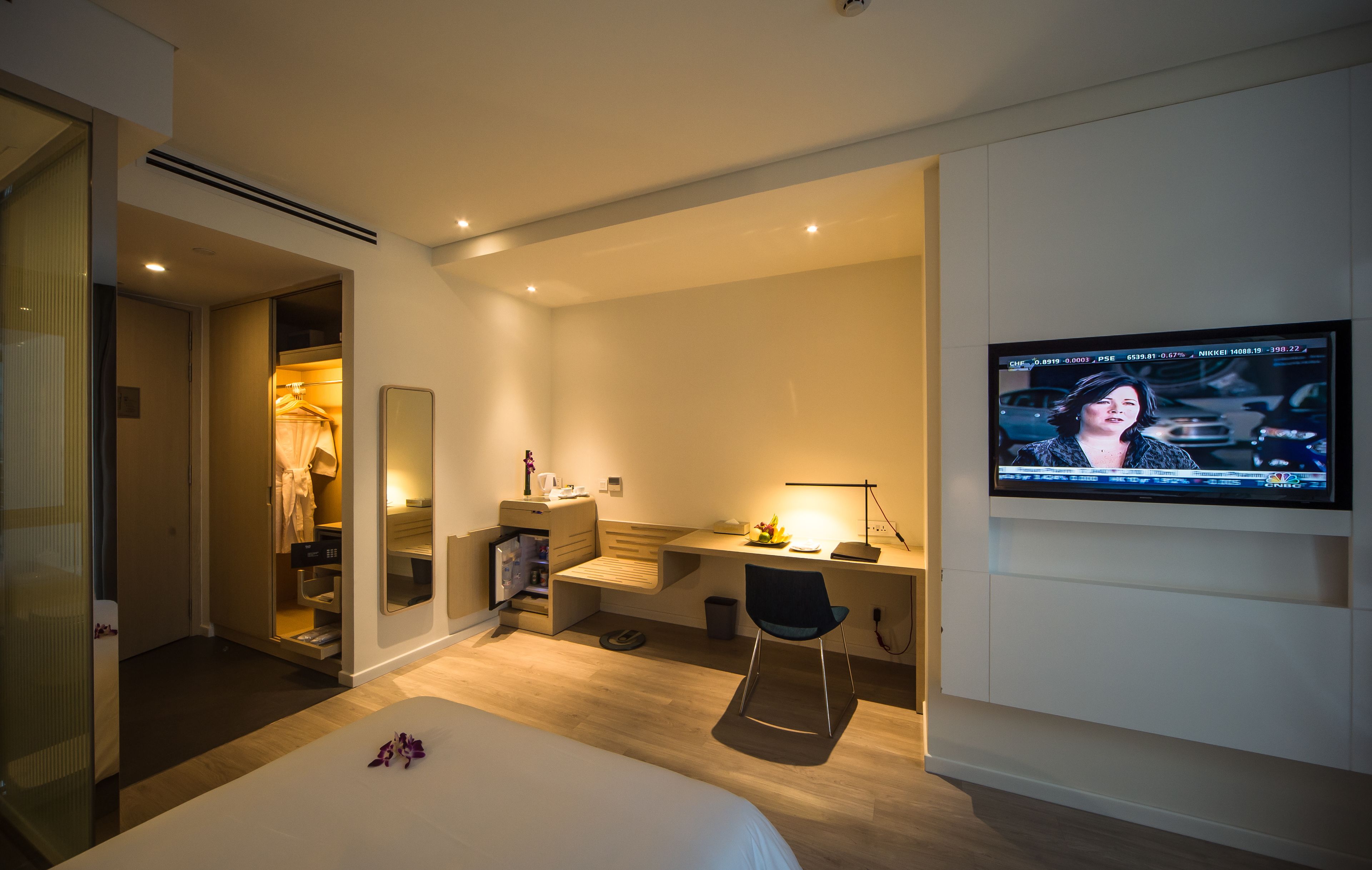 A modern hotel room with a bed, desk, chair, TV, and a walk-in closet.