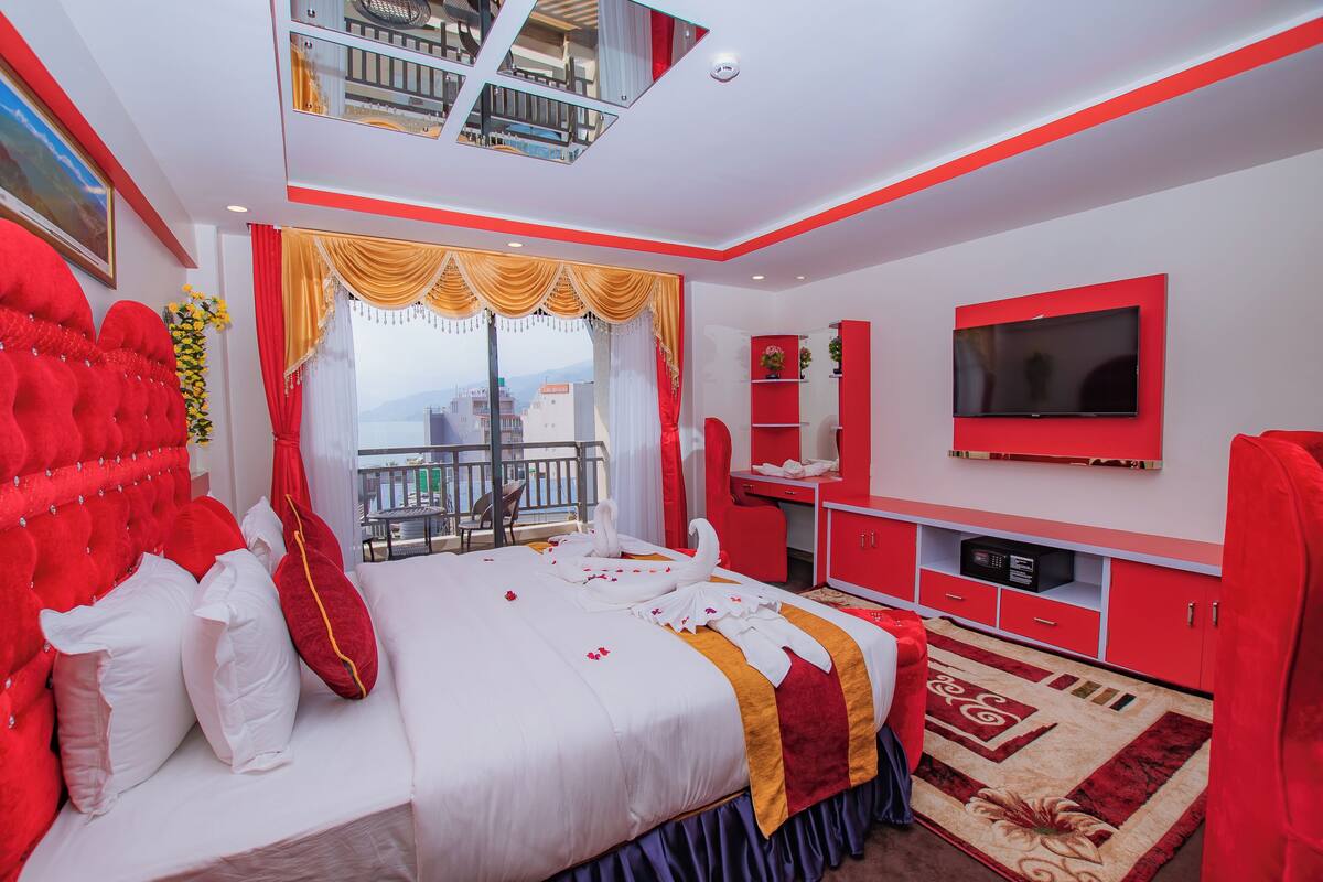 A modern hotel room with a large bed, a balcony with a view, a TV, and red and white decor.