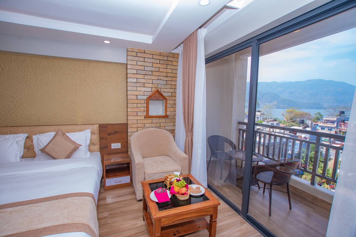 A hotel room with a bed, a chair, a table with fruit and a plate, and a balcony with a view of the mountains.