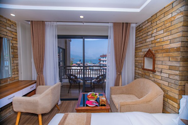 A hotel room with a balcony, a wooden desk, two armchairs, a coffee table with fruit and a book, and a bed. - Hotel Pokhara Village & SPA (Pokhara)