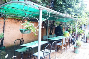 Un café en plein air avec des tables et des chaises disposées sous une pergola verdoyante, entouré de murs en briques et de plantes en pot.