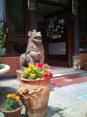 Une statue de lion, avec des fleurs dans un grand pot, un pot plus petit contenant des œillets d’Inde, et l’entrée d’un bâtiment avec une porte en verre et un paillasson rouge.