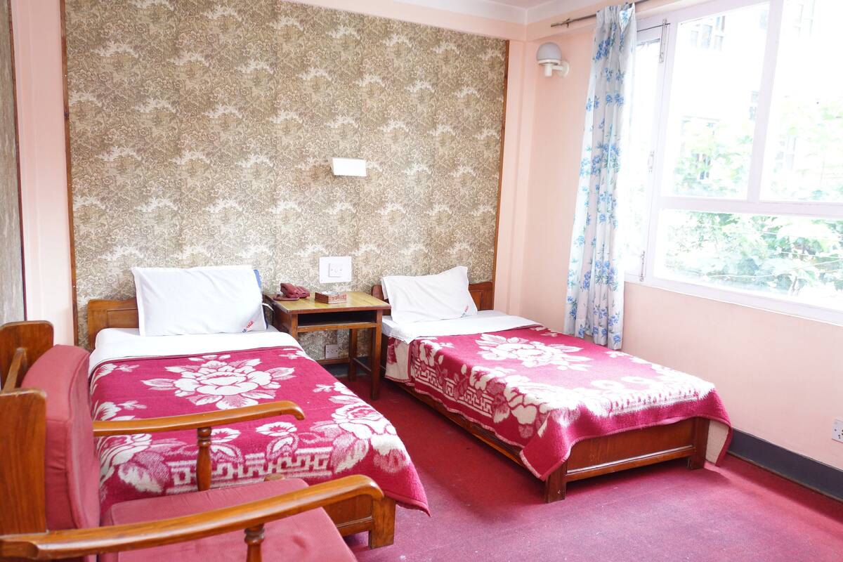 A room with two single beds, a wooden chair, a small table, and a window with curtains.