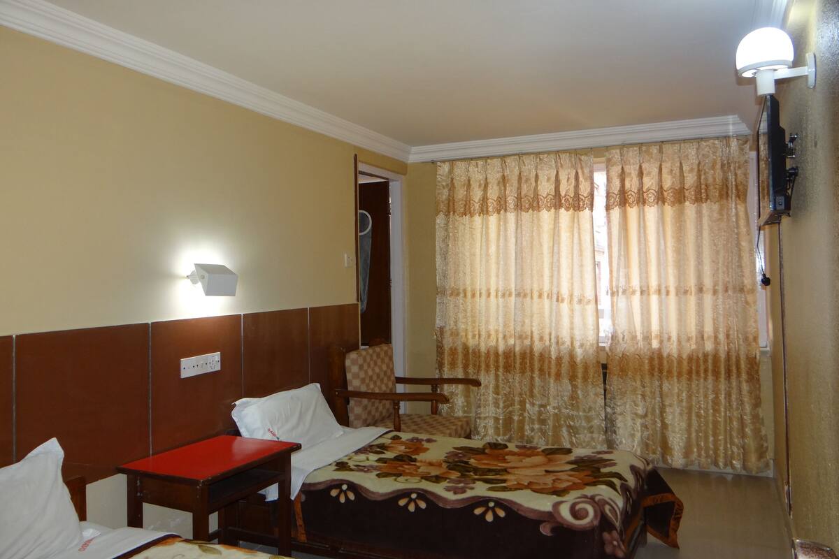 A hotel room with a bed, a desk, a chair, a wall-mounted light, and a window with curtains.