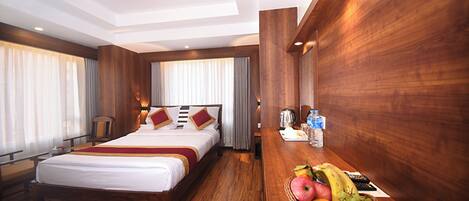 A hotel room with a wooden bed, a desk with a fruit basket, and a view of the city.