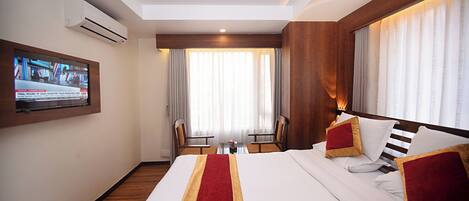 A hotel room with a large bed, a TV mounted on the wall, a wooden floor, and a window with curtains.