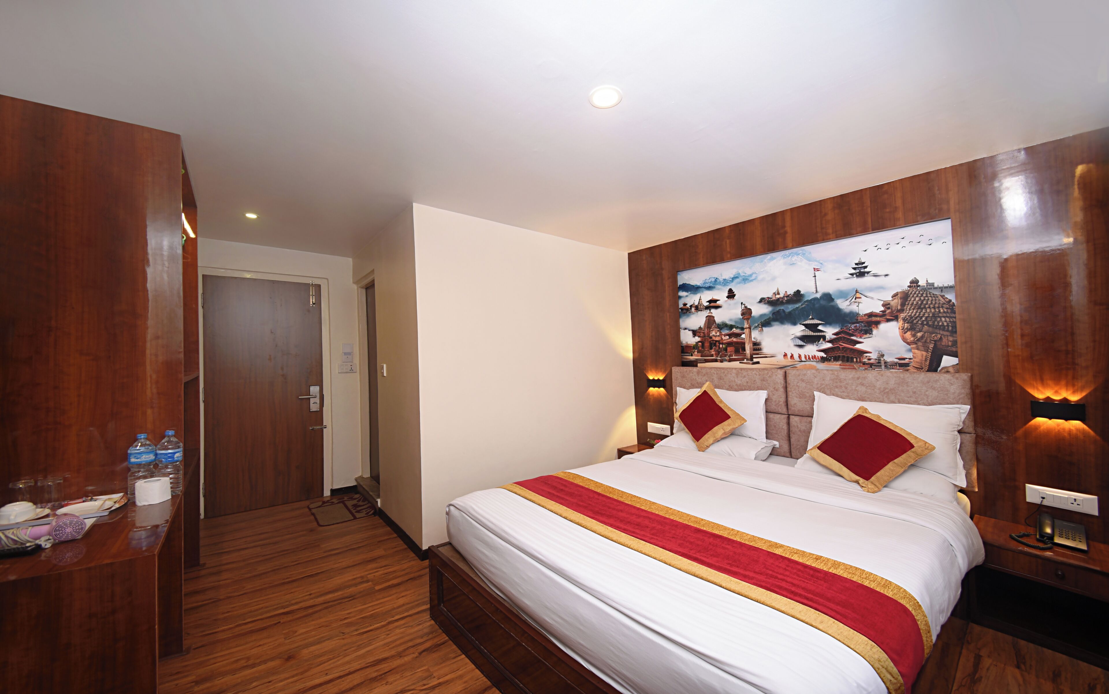 Habitación de hotel con cabecera de madera, cama grande con ropa de cama blanca y detalles en rojo y dorado, un cuadro de una escena tradicional en la pared y un escritorio de madera con artículos de tocador.