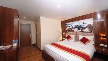 Habitación de hotel con cabecera de madera, cama grande con ropa de cama blanca y detalles en rojo y dorado, un cuadro de una escena tradicional en la pared y un escritorio de madera con artículos de tocador.