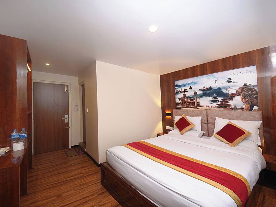 Habitación de hotel con cabecera de madera, cama grande con ropa de cama blanca y detalles en rojo y dorado, un cuadro de una escena tradicional en la pared y un escritorio de madera con artículos de tocador.