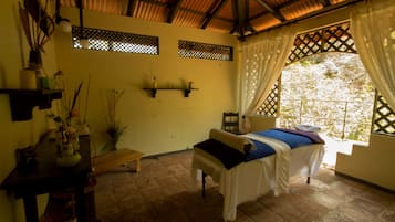 Guest room with a bed, table, and chair, offering a view of lush greenery outside.